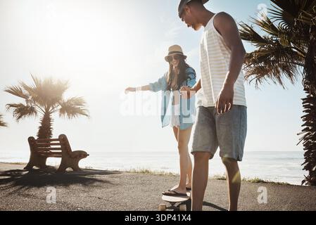 Paare, Skateboard und Hände halten an der Strandpromenade, um zu lernen, glücklich zu sein oder auf der Straße auszubalancieren. Menschen, Mann und Frau mit Raum, interrassische Liebe und Stockfoto