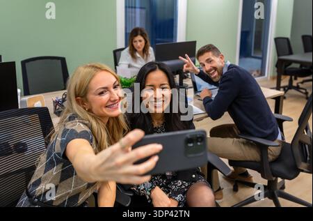 Lächelnde Freundinnen, die ein Büro-Selfie mit der Fotobombe eines Kollegen machen. Kollegen, die Spaß bei der Arbeit haben. Stockfoto