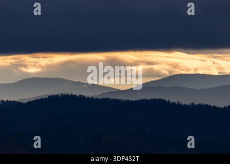 Foto von Schichten nebeliger Bergkämme in Beskiden, Polen, unter einem dramatischen dunklen Himmel mit einem leuchtend goldenen Streifen bei Sonnenuntergang. Stockfoto
