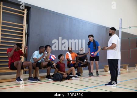 Ein vielfältiges männliches Teenager-Team in Trikots sitzt im Fitnessstudio mit Volleybälle und hört dem Coach beim Halten eines Tablets zu Stockfoto