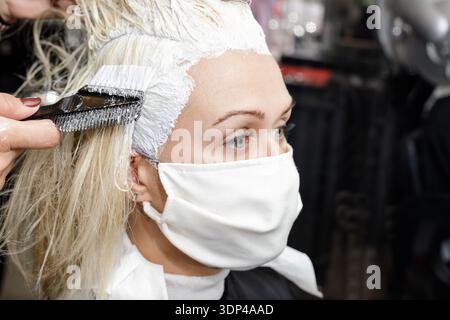 Junge Frau in medizinischer Maske im Schönheitssalon während Haarfärbung und Coronavirus-Pandemie. Neues normales covid19-Konzept Stockfoto