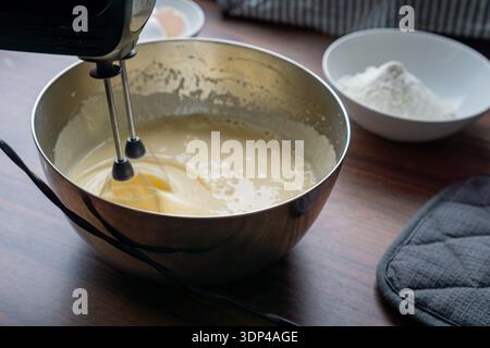 Metallschüssel mit flüssigem Kuchenteig, der von einem elektrischen Handmixer auf einem Holztisch gemischt wird. Stockfoto