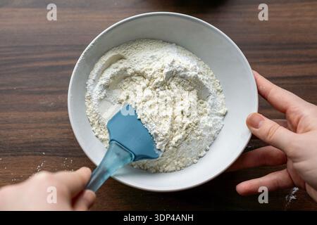 Hände halten eine Schüssel Mehl, während sie mit einem Silikonspachtel umrühren. Die trockenen Zutaten werden auf einem Holztisch zum Backen zubereitet. Stockfoto