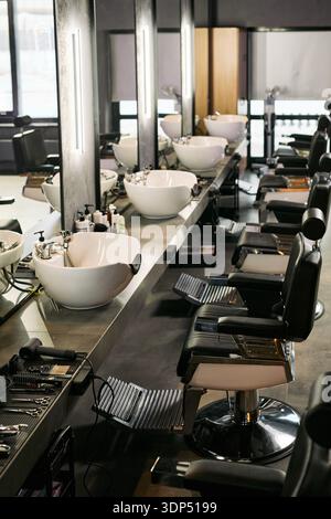 Leerer Friseurladen mit einer Reihe moderner Friseurstühle und Waschbecken, die entlang einer Theke mit verschiedenen Friseurwerkzeugen und Produkten angeordnet sind, die auf den Arbeitsplätzen sichtbar sind Stockfoto
