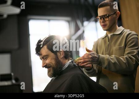 Kaukasischer Mann mittleren Alters sitzt im Barbershop-Stuhl lächelnd, während junger kaukasischer Mann Friseur mit Tattoos Haare mit Kamm und Schere schneidet, Sonnenlicht im Hintergrund scheint Stockfoto