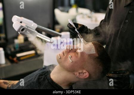Kaukasischer junger erwachsener Mann, der vom Black man Friseur Gesichtsbehandlung erhält, heißes Handtuch und Rasiercreme auf das Gesicht aufträgt, während er im Barbershop-Stuhl mit Dampfmaschine in der Nähe liegt Stockfoto