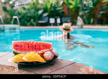 Frischer tropischer Obstteller mit Wassermelone Mangostan Passionsfrucht am luxuriösen Swimmingpool im thailändischen Resort mit einer Frau im Strohhut-Schwi Stockfoto