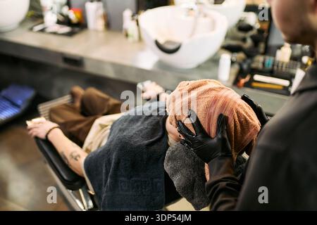 Kaukasischer junger erwachsener Mann, der sich im Barbershop-Stuhl zurücklehnt und heiße Handtuchbehandlungen von schwarzem Mann mittleren Alters erhält, Handschuhe trägt, sichtbares Tattoo auf dem Arm des Kunden, Salonwaschbecken im Hintergrund Stockfoto