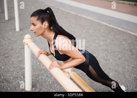 Eine Frau mit starker Passform in sportlicher Kleidung führt eine anspruchsvolle Liegestützübung auf parallelen Außenbars aus und konzentriert sich dabei auf ihr Krafttraining Stockfoto
