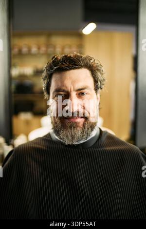 Porträt eines kaukasischen Mannes mittleren Alters, der im Barbershop-Stuhl sitzt und mit beschnittenem Bart und Haar in die Kamera lächelt, mit einem Barber cape, Hintergrund mit verschwommenem Innenraum des Salons Stockfoto