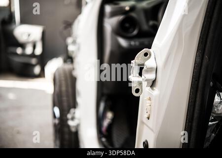 Eine detaillierte Ansicht eines weißen Scharniers der Fahrzeugtür an einer offenen Fahrzeugtür in einer Karosseriebau mit verschwommenem Fahrzeuginnenraum und anderen Teilen im Hintergrund. Stockfoto