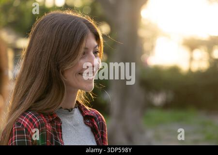 Erwachsene Frau lächelt, sieht rechts aus und steht im Park mit kariertem Hemd, grauem Oberteil und Choker Stockfoto