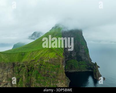 Blick aus der Vogelperspektive auf den Leuchtturm von Kallur, der dramatisch auf einer grünen Klippe thront, umgeben von Nebel und überragt das dunkle, wirbelnde Meer, Kalsoy, Stockfoto