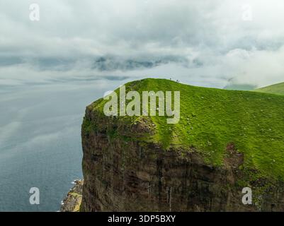 Aus der Vogelperspektive auf die smaragdgrüne Klippe, die das tiefblaue Meer unter einer Decke aus brodelnden Wolken trifft, Kalsoy, Färöer Inseln. Stockfoto