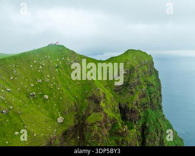 Aus der Vogelperspektive des Leuchtturms von Kallur, der Wache auf den dramatischen, grünen Klippen steht, wo das Land unter einem riesigen, bewölkten Himmel auf das Meer trifft, Kalsoy, Stockfoto