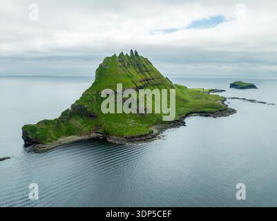 Aus der Vogelperspektive erhebt sich die scharfe, smaragdgrüne Insel Tindhólmur dramatisch aus dem stahligen Meer, deren zerklüftete Gipfel den nebeligen Himmel durchdringen, Tindholmur, Färöer Stockfoto