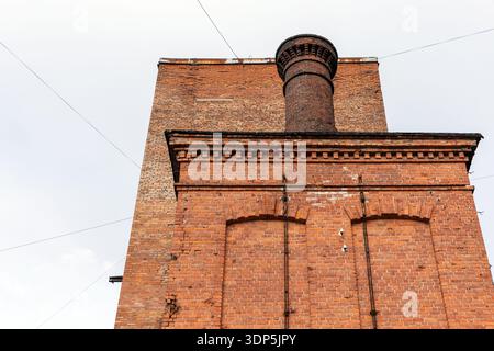 Markanter roter Backsteinkamin erhebt sich über einer historischen Fabrikfassade und zeigt Industriearchitektur, verwittertes Mauerwerk und urbanes Erbe Stockfoto