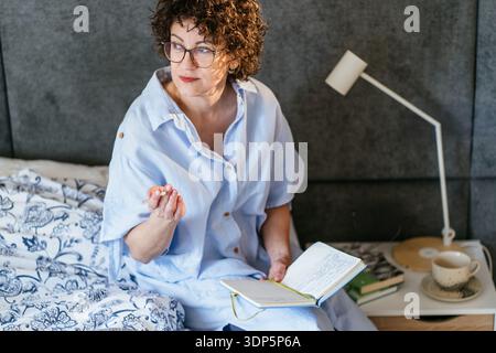 Eine nachdenkliche weiße Frau in ihren 40ern mit Brille, die im Schlafzimmer das Journalieren anhält Stockfoto