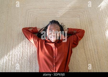 Optimistische, fröhliche Afroamerikanerin, die zu Hause auf Jute-Teppich liegt und Musik in Schoten hört. Stockfoto