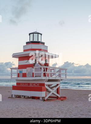 Ein lebendiger Rettungsschwimmer-Turm hebt sich am Miami Beach ab, während die Sonne untergeht, während ein Paar einen romantischen Moment teilt. Die sanften Wellen und der pastellfarbene Himmel schaffen eine ruhige Atmosphäre für Strandgäste. Stockfoto
