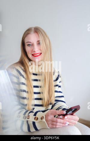 Glückliche junge Frau, die auf dem Handy schreibt. Digitaler Lifestyle, Fernkommunikation und lässiges Porträt in Innenräumen. Stockfoto