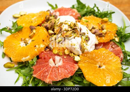 Nahaufnahme eines Zitrussalats mit Orangen- und Grapefruitscheiben, garniert mit Sahne und gehackten Pistazien auf frischem Rucola-Grün auf einer weißen Platte. Stockfoto