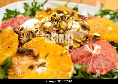 Nahaufnahme eines frischen Zitrussalats mit Orangen- und Grapefruitscheiben, garniert mit Sahne und gehackten Pistazien auf einem Bett aus Blattgemüse. Stockfoto