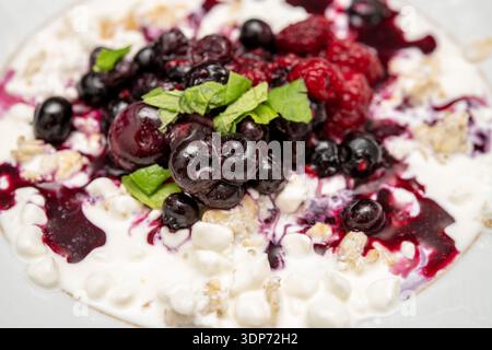 Nahaufnahme von Hüttenkäse mit gemischten Beeren, Minzblättern und Beerensauce auf einem weißen Teller in einer hellen Umgebung. Stockfoto