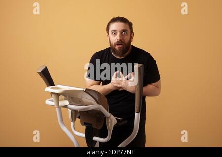 Bärtiger Mann in schwarzem T-Shirt, der überrascht aussieht, während er auf dem Heimtrainer vor beigefarbenem Hintergrund steht. Stockfoto