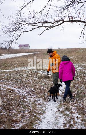 Ein hübsches Paar schlendert Hand in Hand durch eine malerische Winterlandschaft, begleitet von ihrem verspielten Hund auf einem verschneiten Pfad, umgeben von Ruhe Stockfoto