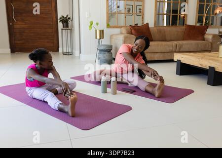 Afroamerikanische Mutter und Kind dehnen sich in Aktivkleidung auf lila Matten zu Hause mit Flaschen, Telefon Stockfoto
