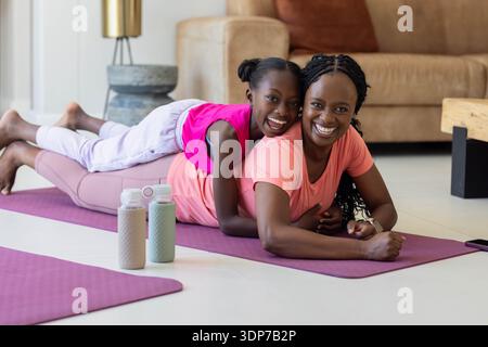Afroamerikanische Mutter und Tochter in Aktivkleidung, spielen auf lila Matten, Flaschen im Wohnzimmer Stockfoto