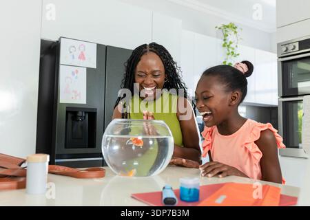 Afroamerikanische Mutter und Kind lachen und lehnen sich über Fischschüssel mit Orangengoldfisch in der Küche Stockfoto