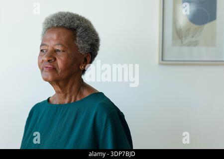 Ältere afrikanische Frau, die im schlichten Zimmer mit blaugrünen Nieten und gerahmten Kunstwerken, Kopierraum, zurückblickt Stockfoto