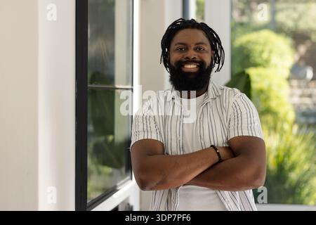Afrikanischer Mann, der Arme im Eingang durch schwarz gerahmtes Fenster überkreuzt, gestreiftes Hemd, perlenbesetztes Armband Stockfoto