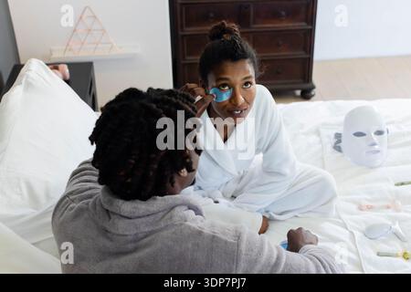 Afrikanisches Paar, das Bademäntel trägt, sitzt im Schlafzimmer auf dem Bett und trägt blaues Hydrogel-Patch an Stockfoto