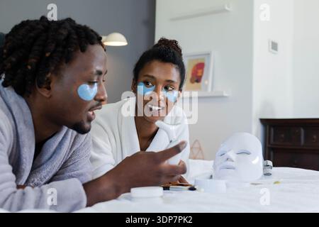 Afroamerikanisches Paar im Bett, trägt Bademäntel und Augenflecken, Mann hält Gerät, Frau beobachtet Stockfoto