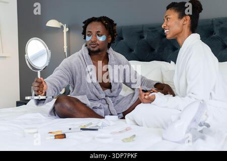 Afroamerikanisches Paar, das in Bademänteln auf dem Bett sitzt und einen Spiegel hält Stockfoto