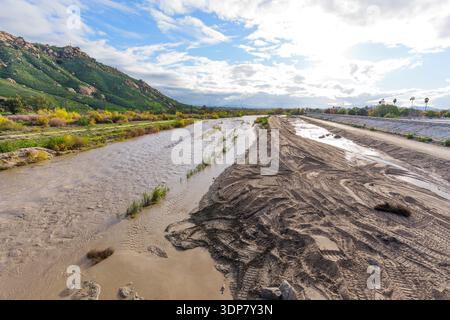 Schlammiges Wasser fließt durch das Flussbett, Reifenspuren auf Sand, den grünen Mount Rubidoux und den Betondeich unter hellem, bewölktem Himmel. Stockfoto