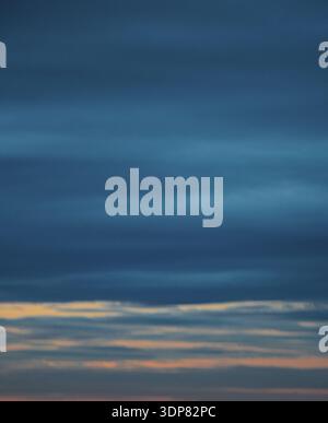 Dramatische vertikale Aufnahme mit schweren dunkelblauen Wolken, die die obere Atmosphäre mit einem dünnen goldenen Licht am Horizont einnehmen. Stockfoto