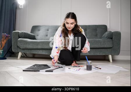 Frau malt mit Farben und Pinseln, während sie drinnen auf dem Boden sitzt. Der Künstler steckt in Kreativität und kreiert zu Hause farbenfrohe Aquarellmalereien. Stockfoto