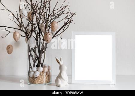 Osterkomposition mit dekorativen Eiern auf Zweigen in Glasvase und leerem Fotorahmen für saisonale Begrüßungsgestaltung mit Kopierraum. Stockfoto