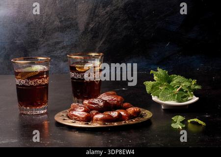 Datteln und zwei Gläser erfrischenden Minztee mit frischen Minzblättern sind auf einem dunklen Tisch angeordnet. Darstellung traditioneller Elemente für schnelle Dur Stockfoto