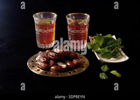 Traditioneller marokkanischer Pfefferminztee in kunstvollen Gläsern mit frischen Datteln auf einem goldenen Teller und Minzzweigen, symbolisiert iftar und den Bruch des schnellen Duris Stockfoto
