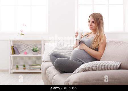 Schwangere Frau sitzt auf der Couch und trinkt Milch im hellen Wohnzimmer Stockfoto