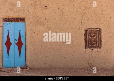 140 geschlossene blaue Tür und ein gegrilltes Fenster eines traditionellen, von lehmwänden umgebenen Amazigh-Hauses (Berber) in Khamlien, neben Erg Chebbi. Merzouga-Marokko Stockfoto