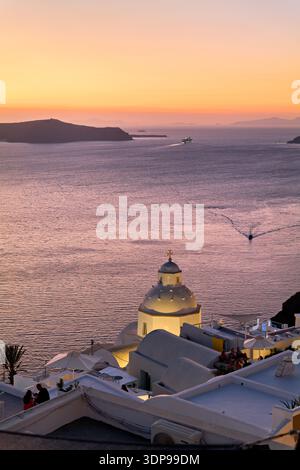 Ein ruhiger Sonnenuntergang in Santorin, Griechenland, beleuchtet eine Kirchenkuppel und Caldera, während die Menschen auf Dächern entspannen und Boote die Ägäis kreuzen. Stockfoto