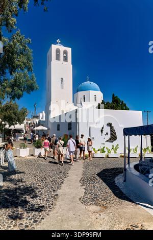 Touristen erkunden eine bezaubernde Kopfsteinpflasterstraße in Santorin, Griechenland, vorbei an berühmten weißen Gebäuden mit blauen Kuppelkirchen unter klarem Himmel. Stockfoto