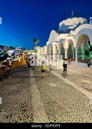 Touristen und Einheimische genießen einen abendlichen Spaziergang auf einer kopfsteingepflasterten Straße in Fira, Santorin, Griechenland, und bewundern die beleuchtete weiße Kirche und die Klippe Stockfoto