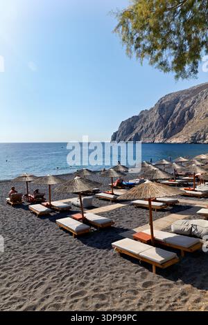 Touristen entspannen sich auf Sonnenliegen unter Strohschirmen an einem schwarzen Sandstrand in Santorin, Griechenland, und genießen das sonnige Wetter und das ruhige Meer. Stockfoto